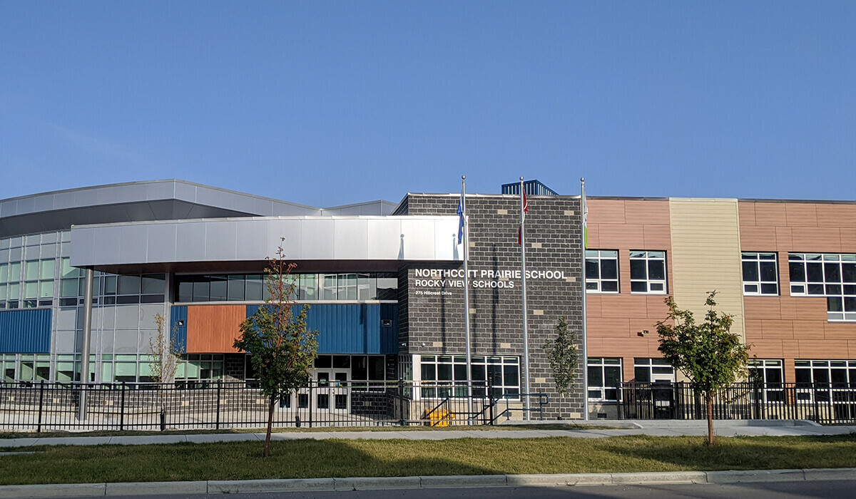 Picture of the front of Northcott Prairie School. Green leafy trees in front of the school are small and beginning to grow. Blue sky above.